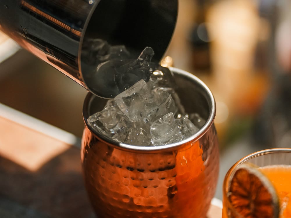 A bartender tipping ice from a shaker into a copper mug beside an orange cocktail