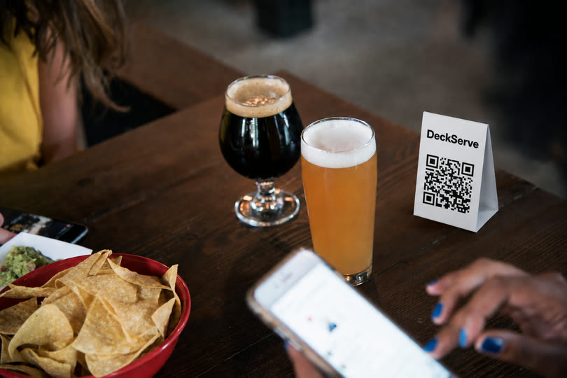 A pub table with beers, snacks and a DeckServe QR card while a guest orders from their phone